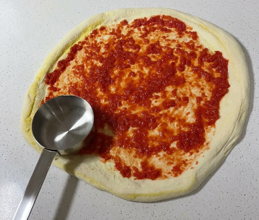 Un mestolo di metallo che distribuisce la salsa di pomodoro sul panetto di pizza steso.
