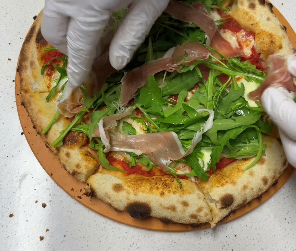 Fette sottili di prosciutto crudo adagiate sopra il letto di rucola sulla pizza.