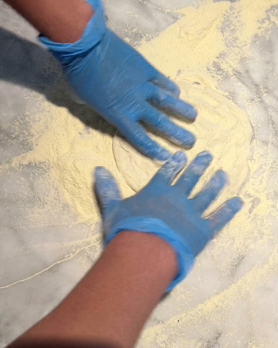 Mani con guanti blu che premono un panetto rotondo di impasto per pizza su un letto di semola su un piano di marmo.
