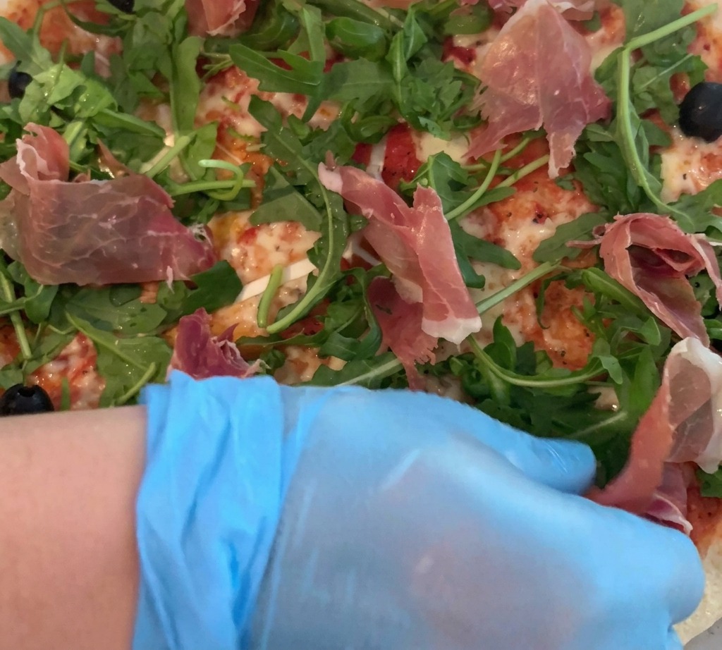 Una mano con un guanto che adagia con cura sottili fette di prosciutto crudo sopra una pizza guarnita con rucola fresca.