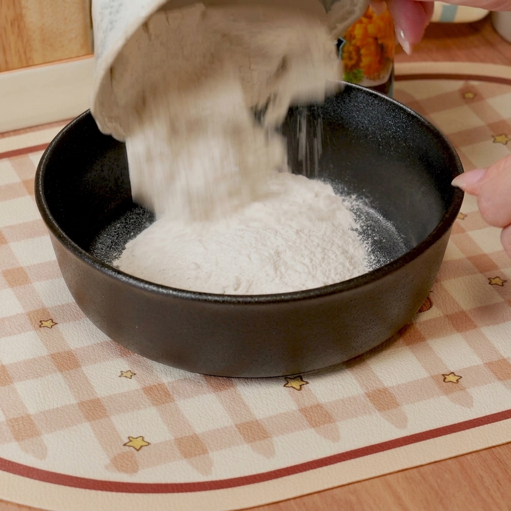 Una frusta che mescola farina per tempura e acqua in una ciotola scura creando una pastella liscia e fluida.