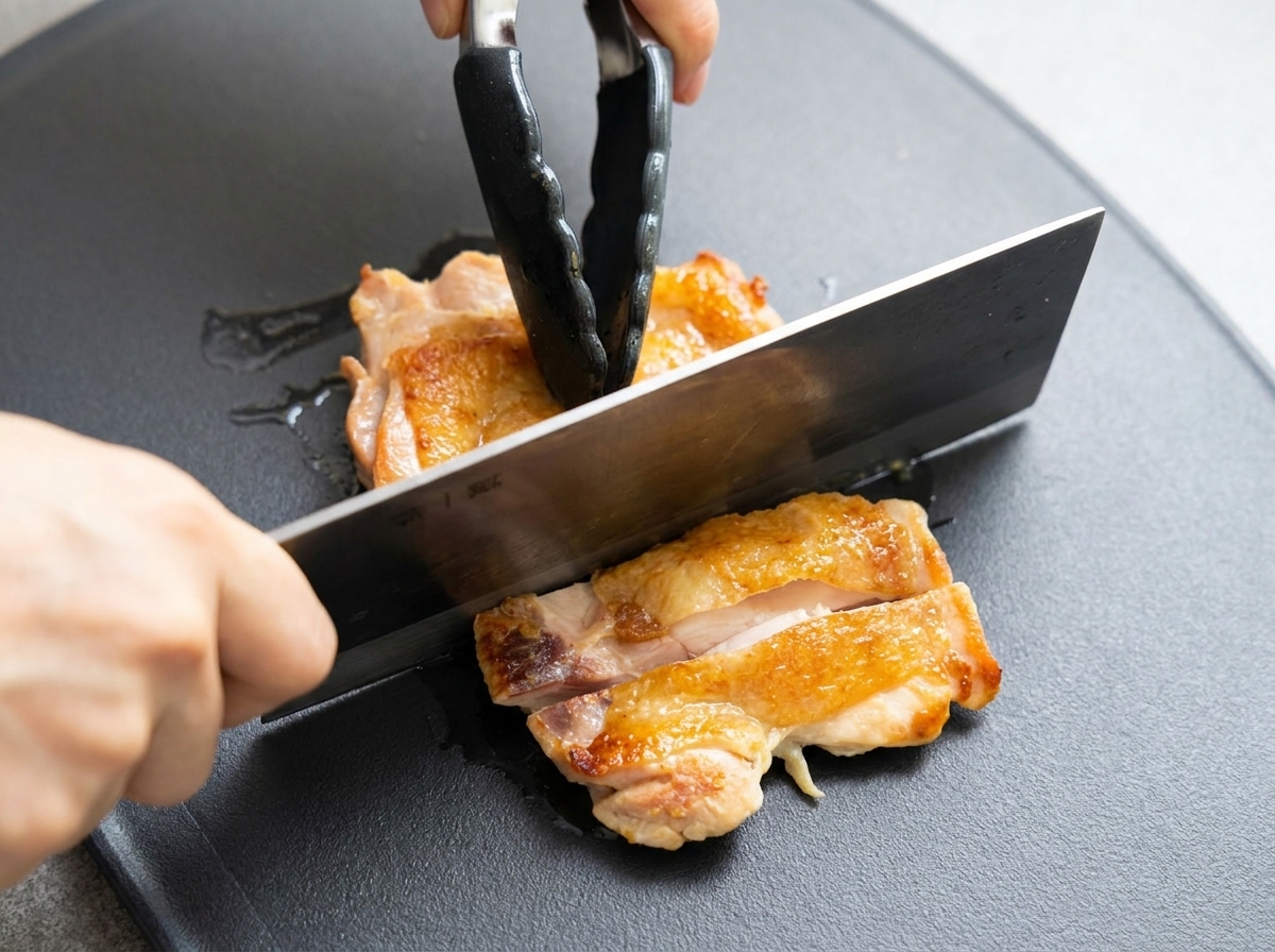 Coscia di pollo dorata tagliata a pezzi su un tagliere scuro con una mannaia.
