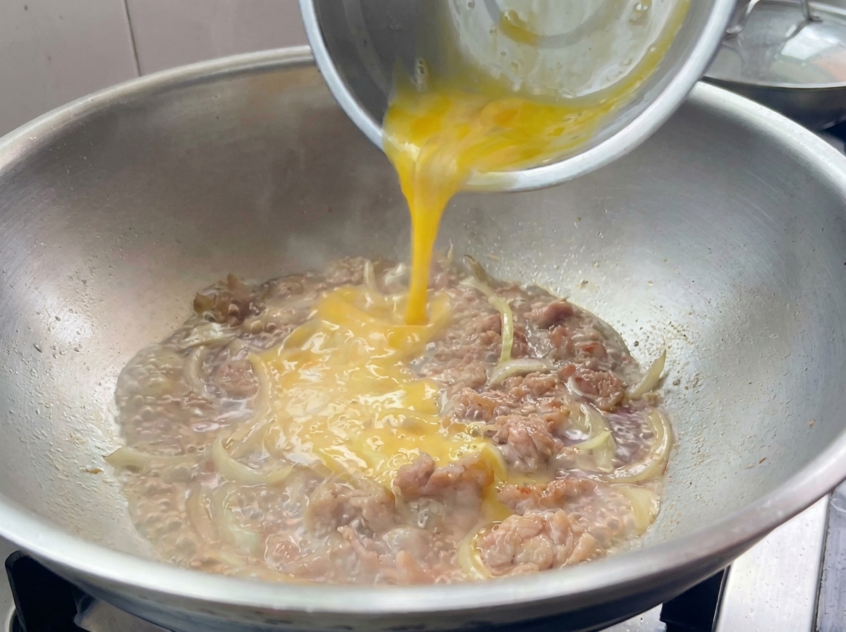 Miscela di uova sbattute versata sopra il maiale e le cipolle in padella.