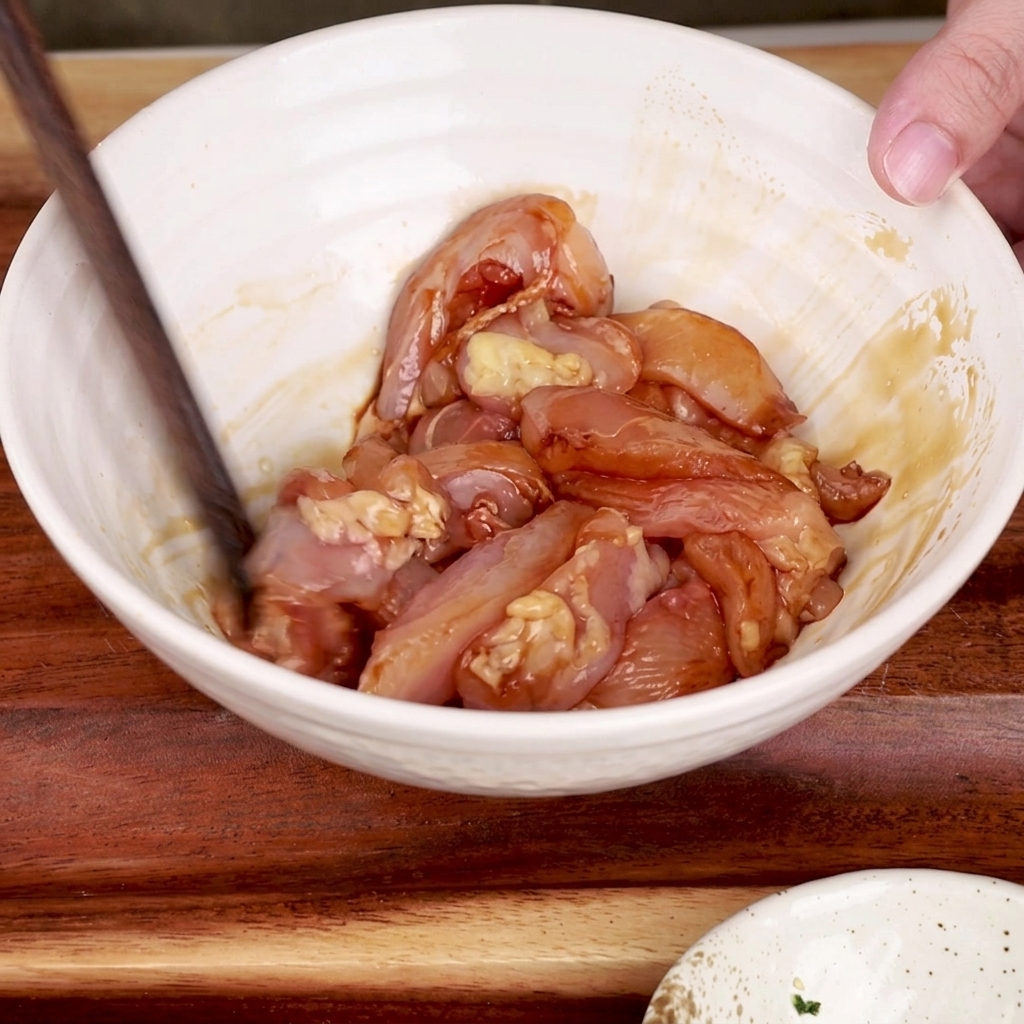 Un paio di bacchette di legno che mescolano i bocconcini di pollo marinati in una ciotola bianca.