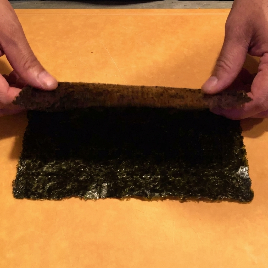 Mani che utilizzano forbici da cucina per tagliare un foglio di alga nori in strisce sottili sopra una ciotola.