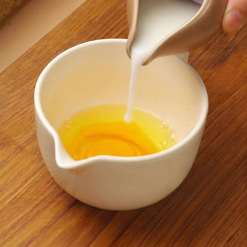 Latte bianco versato da una piccola brocca in un misurino bianco contenente uova crude sbattute.