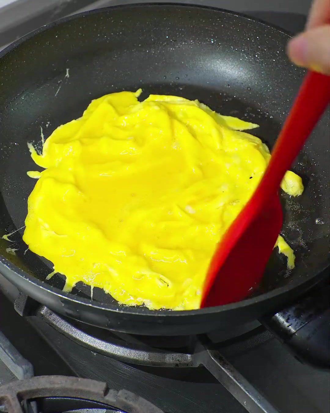Una spatola rossa piega un uovo strapazzato giallo brillante e parzialmente cotto in una padella nera.