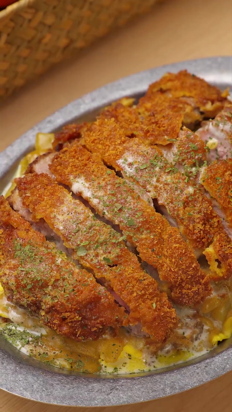 Croccante cotoletta di maiale a fette servita su uova strapazzate morbide e riso, guarnita con curry al cocco e una golosa colata di formaggio fuso.