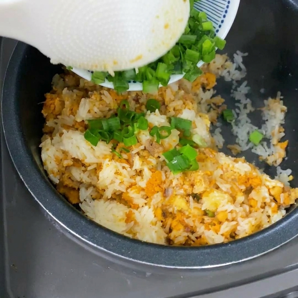 Un cucchiaio bianco che versa cipollotti freschi su riso dal colore dorato nella pentola del cuociriso.