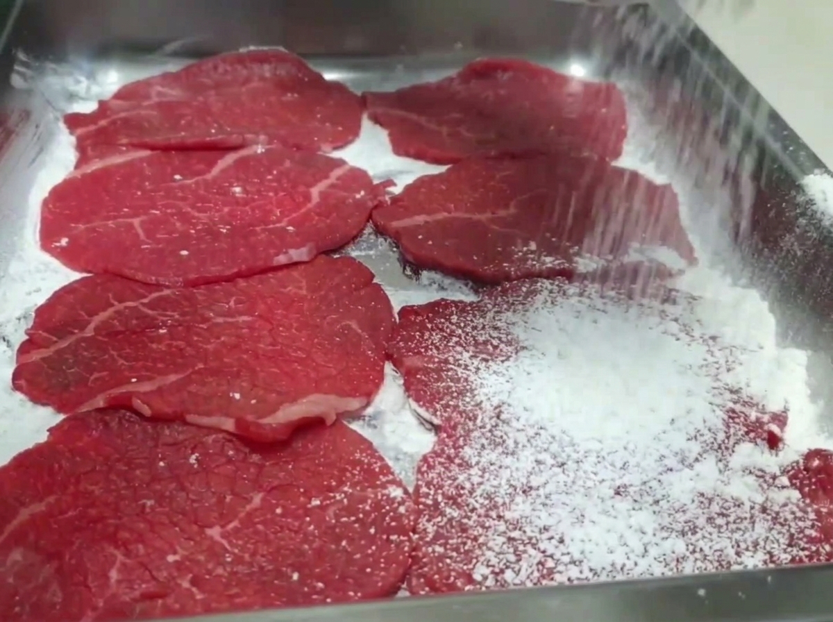 Mano che spolvera farina bianca sulle fette di manzo crudo disposte in un vassoio.