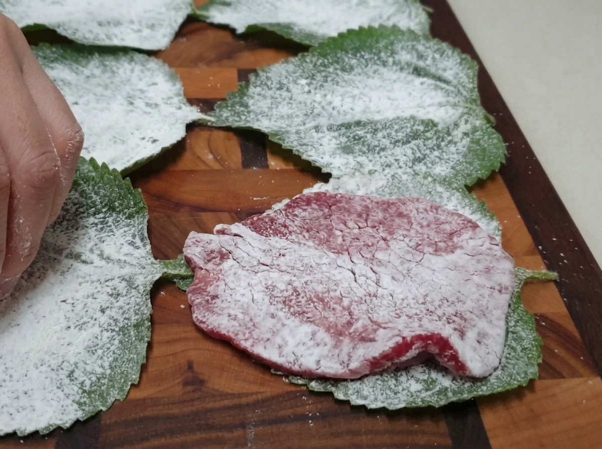 Una fetta di manzo infarinata posizionata sopra una foglia di perilla verde anchessa infarinata, su un tagliere di legno.