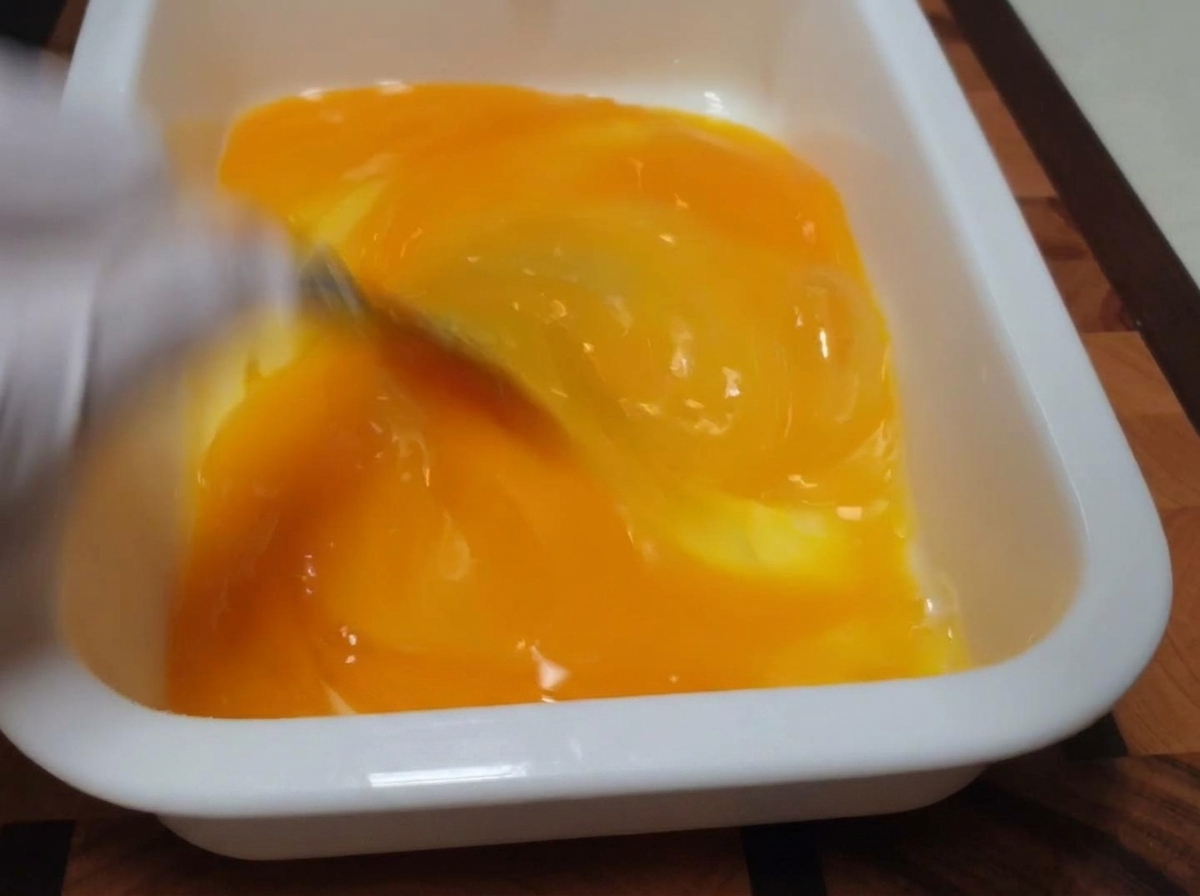 Una frusta che mescola tuorli e albumi di un giallo brillante allinterno di un vassoio bianco rettangolare.