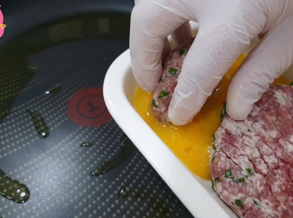 Una mano guantata che immerge un pezzo di manzo e foglia di perilla in un piatto con pastella duovo dorata.