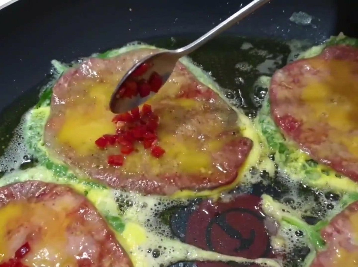 Primo piano di un cucchiaio che aggiunge peperoncini verdi e rossi a dadini sopra le frittelle di manzo in cottura in padella.