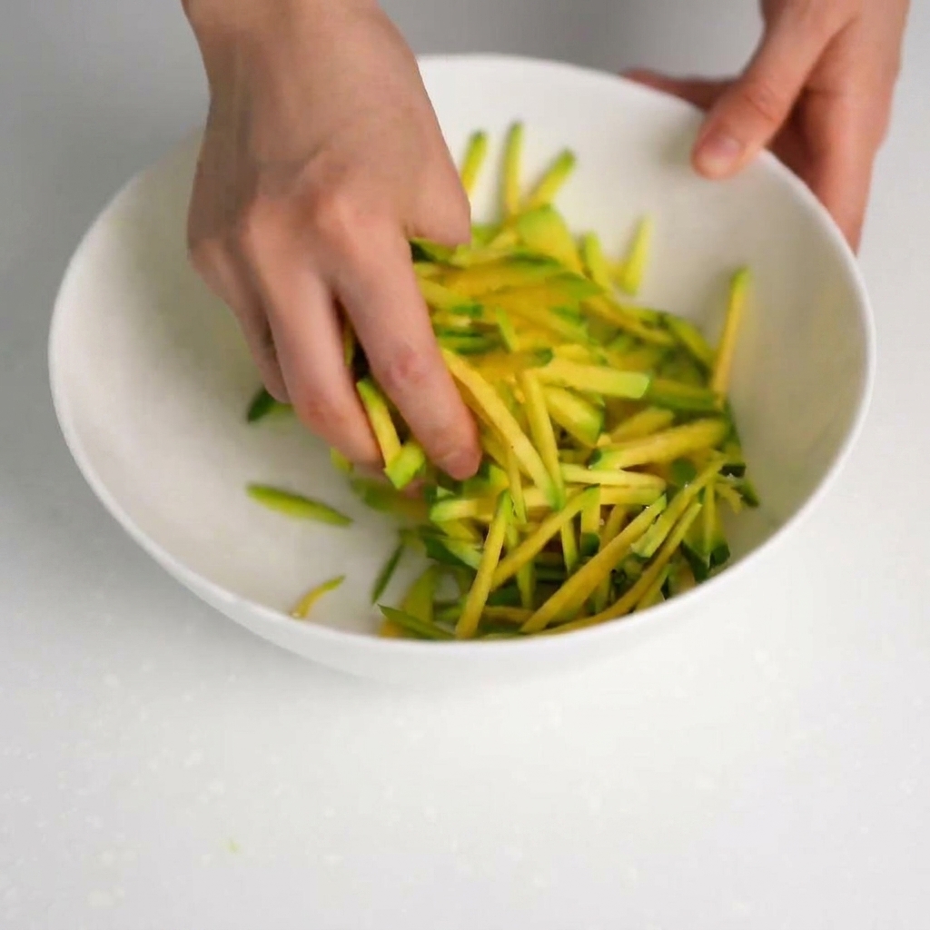 Mani che premono le strisce di zucchine salate per scolare il liquido in una ciotola pulita.