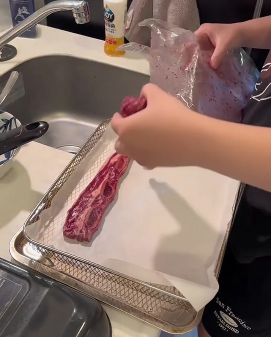 Costine di manzo crude disposte ordinatamente su un foglio di carta da forno allinterno del cestello di una friggitrice ad aria.