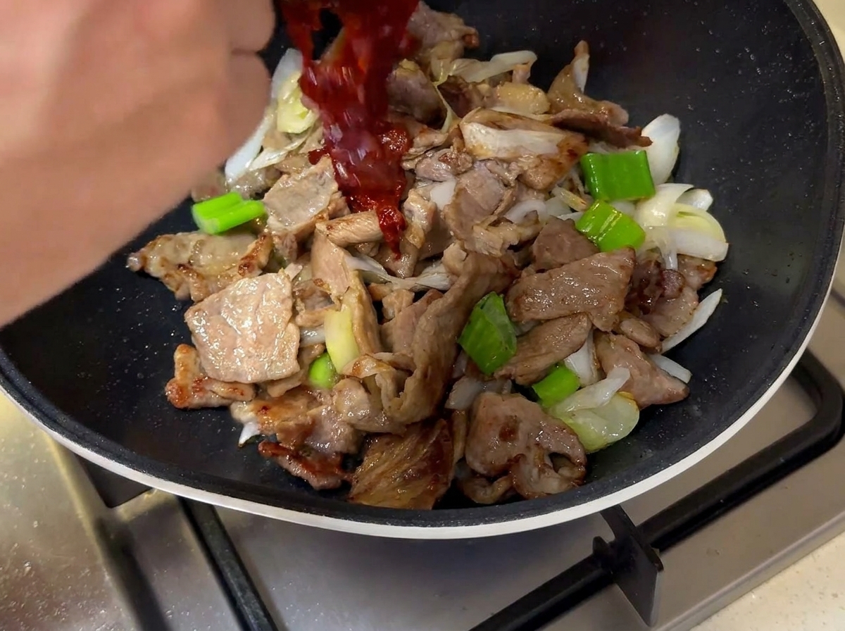 La salsa rosso scuro versata da una ciotolina nella padella sopra la carne e i cipollotti.