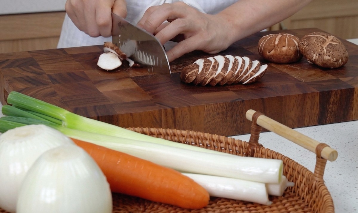 Mani che usano un grande coltello da cucina per affettare funghi shiitake freschi su un tagliere di legno.