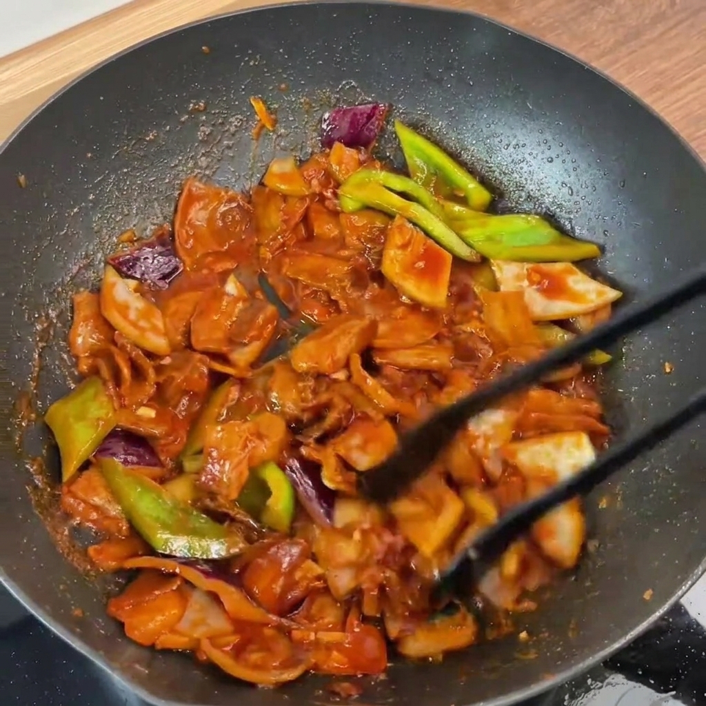 La pancetta di maiale e le verdure saltate in padella vengono mescolate insieme con delle pinze, perfettamente ricoperte dalla salsa rossa lucida.