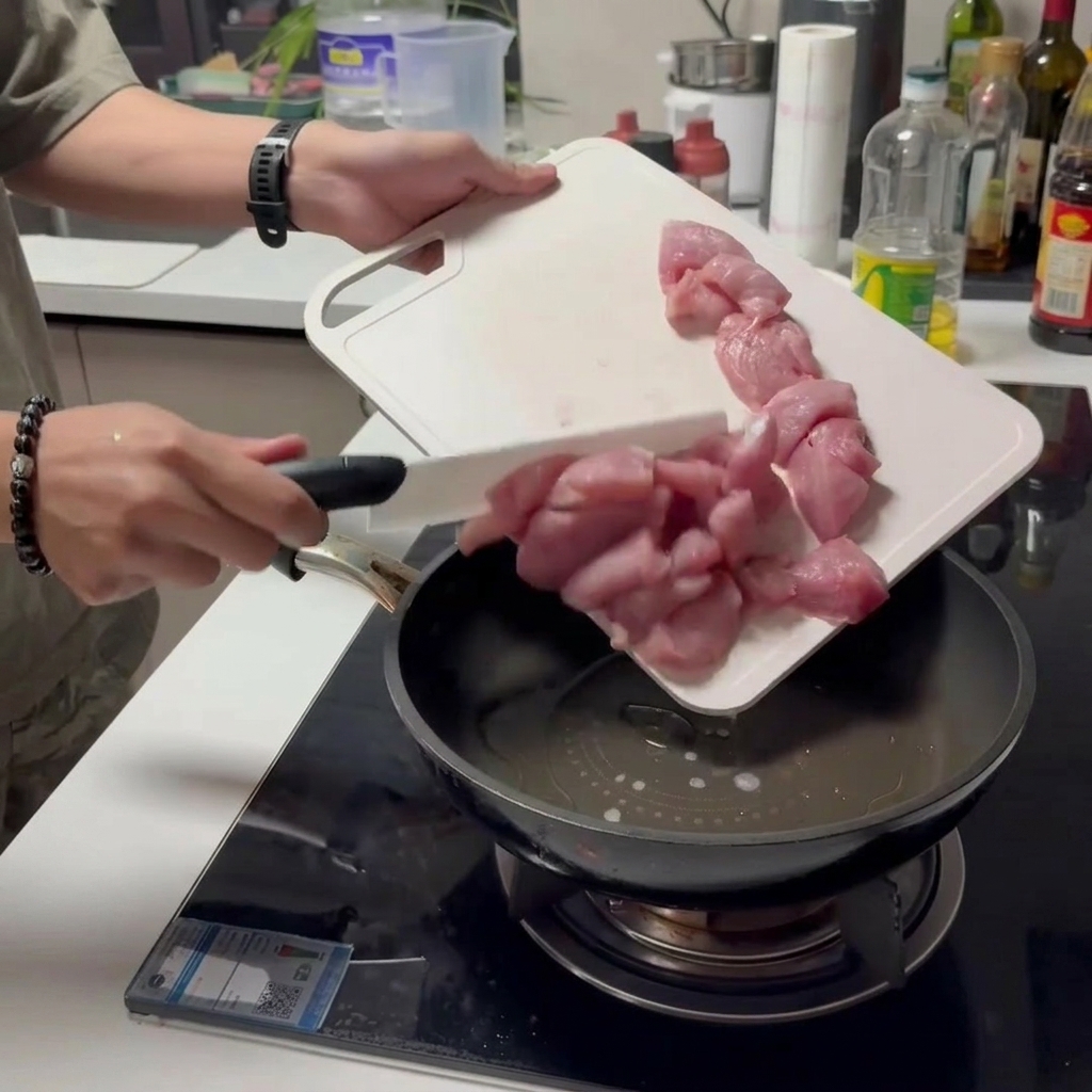 Pezzi di maiale crudo fatti scivolare da un tagliere bianco direttamente in una padella oliata.
