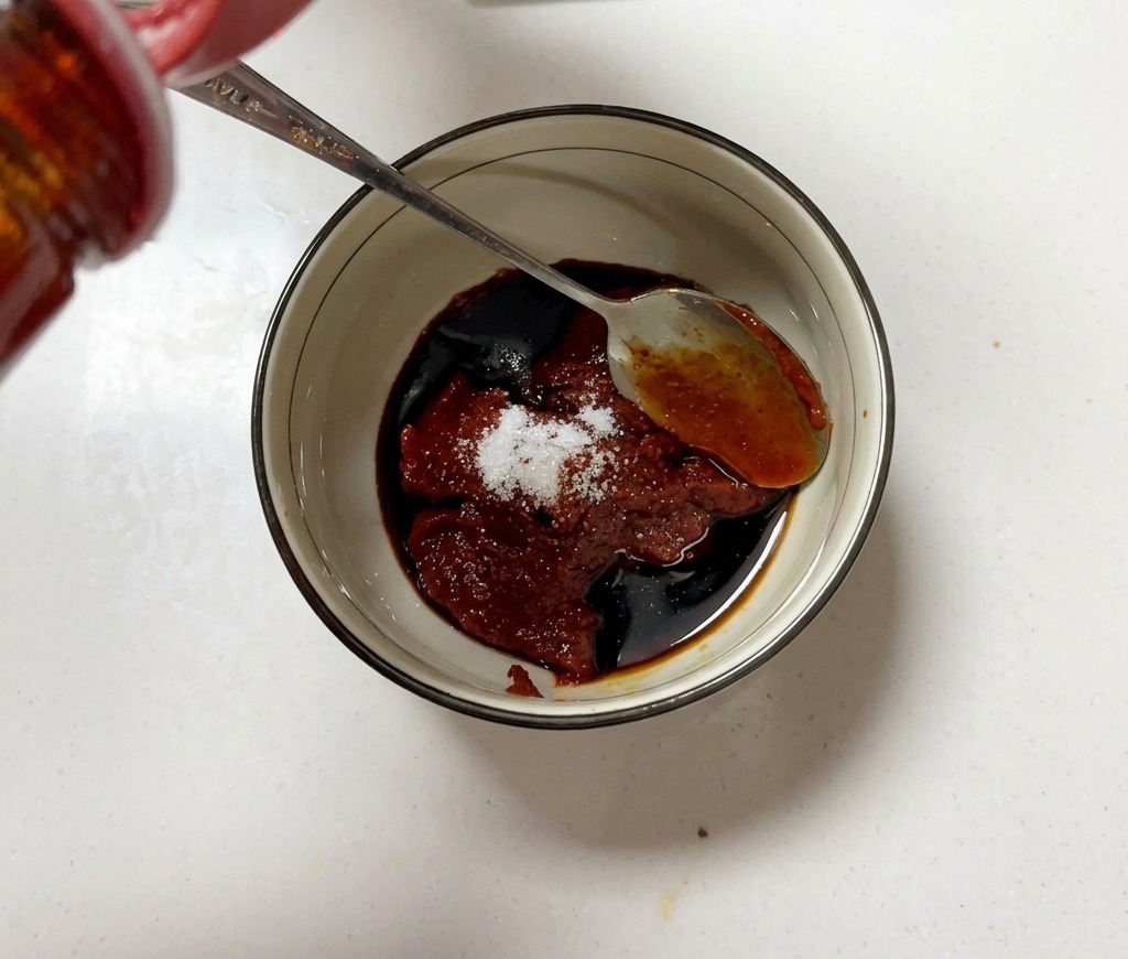 Una piccola ciotola riempita con pasta di peperoncino, zucchero e salsa di soia mentre viene mescolata con un cucchiaio di metallo.