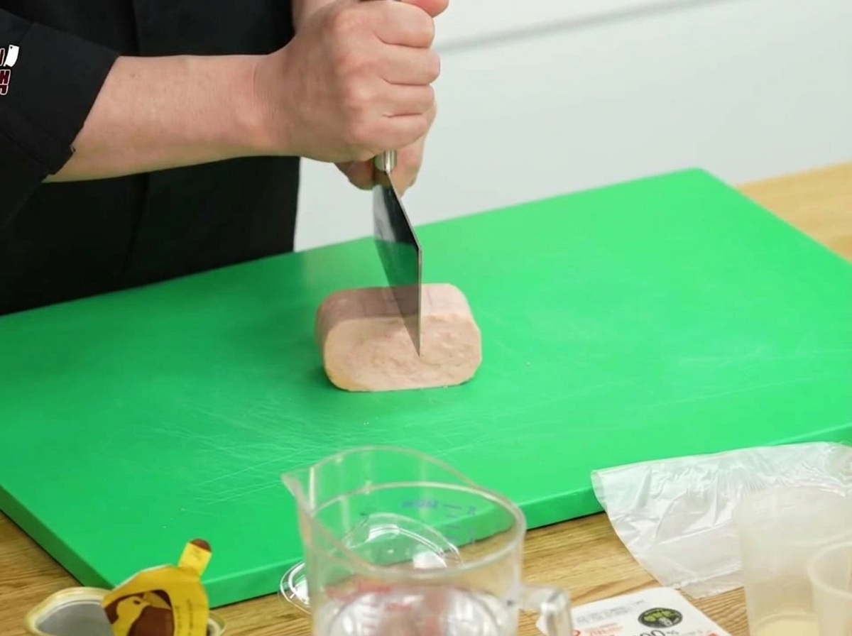 Un blocco di Spam tagliato a metà su un tagliere verde con un grande coltello da cucina.