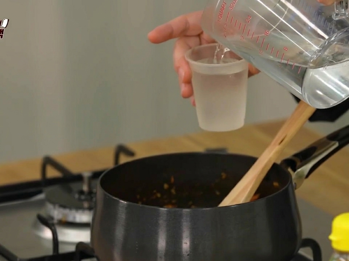 Acqua versata da un misurino in una pentola nera contenente gli ingredienti saltati.