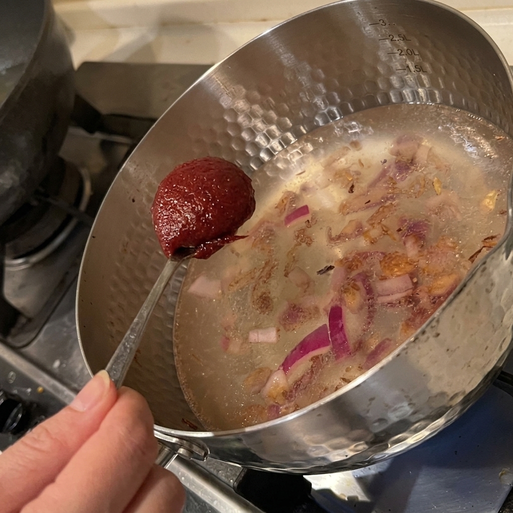 Un cucchiaio di pasta di peperoncino coreana aggiunto in una pentola dacqua con cipolle soffritte.
