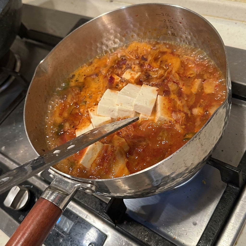 Una varietà di ingredienti freschi, tra cui cubetti di tofu, funghi enoki e kimchi, che sobbollono in un brodo rosso piccante.