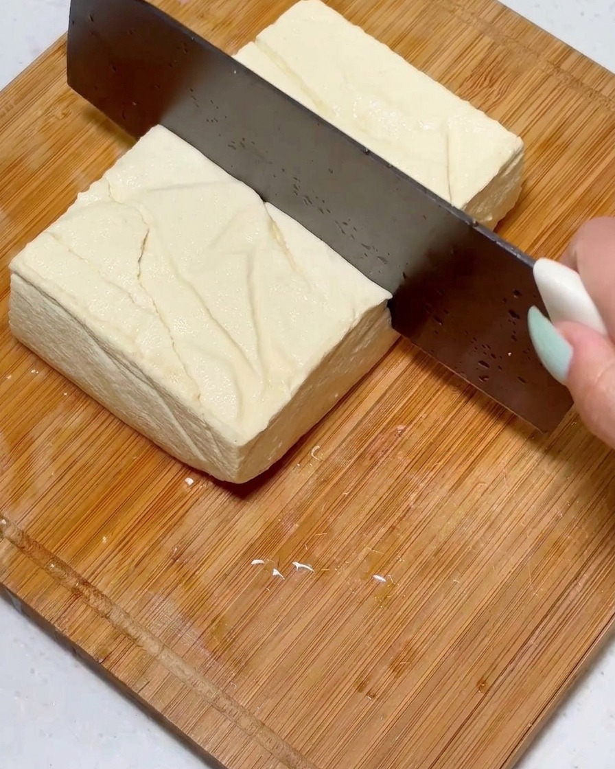 Affettare un blocco quadrato di tofu sodo in pezzi uniformi usando un coltello grande su una superficie di legno.