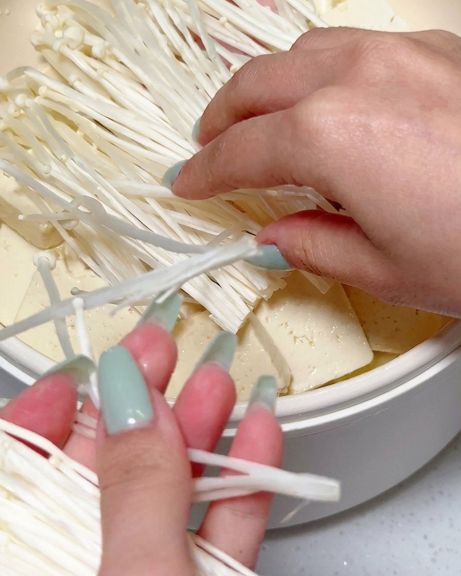 Mani che posizionano un grappolo di funghi enoki bianchi freschi sopra il tofu affettato e altri ingredienti in una pentola.