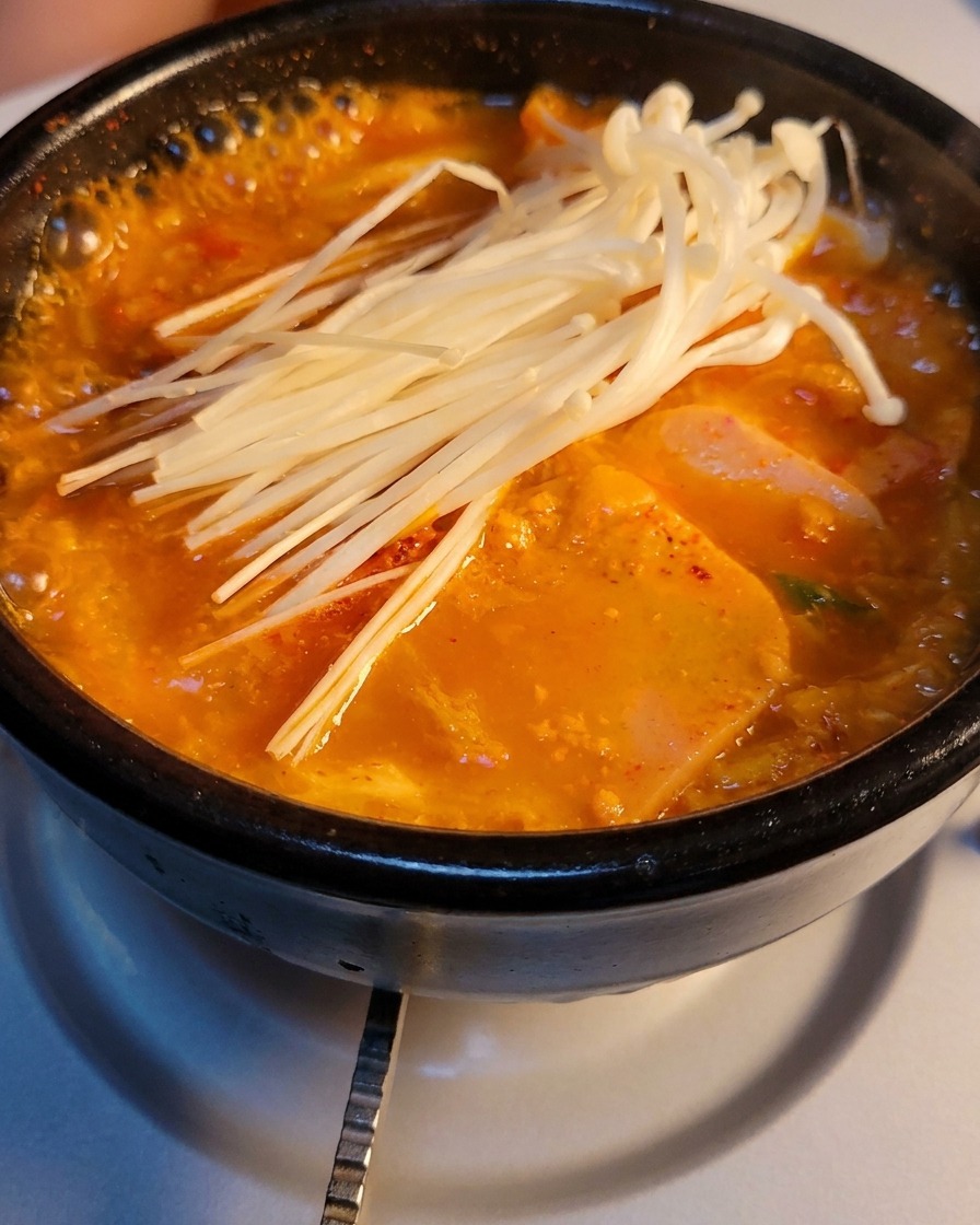 Una pentola di pietra di zuppa gorgogliante di kimchi e carne in scatola guarnita con un mazzetto di funghi enoki freschi.