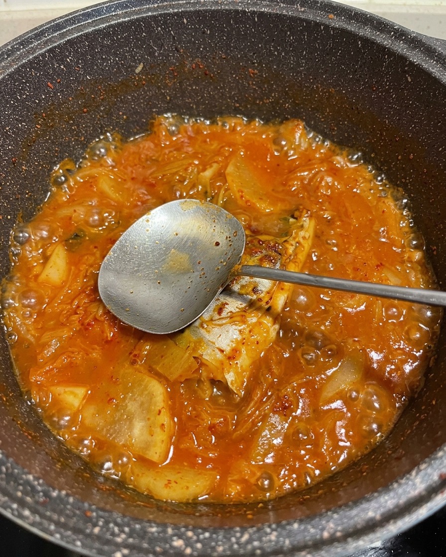 Uno stufato rosso, vivace e gorgogliante di sgombro e verdure in una pentola, con un cucchiaio di metallo che solleva un pezzo di pesce.