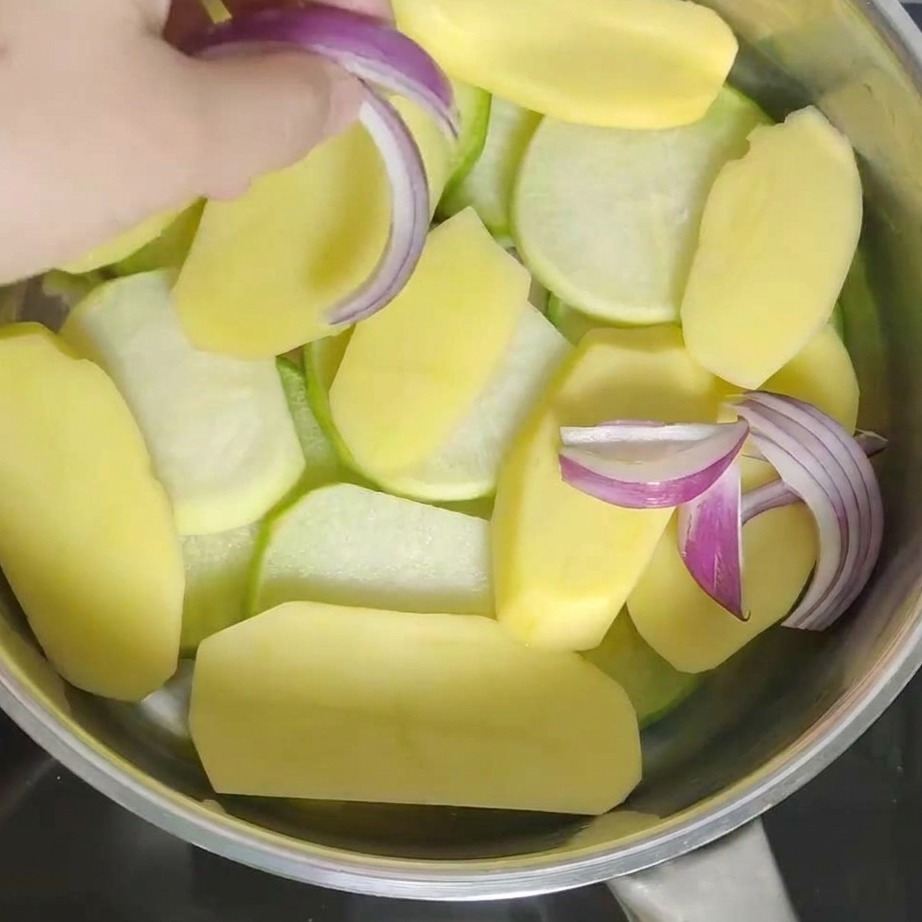 Mano che aggiunge pezzi di cipolla rossa fresca nella pentola sopra le patate.
