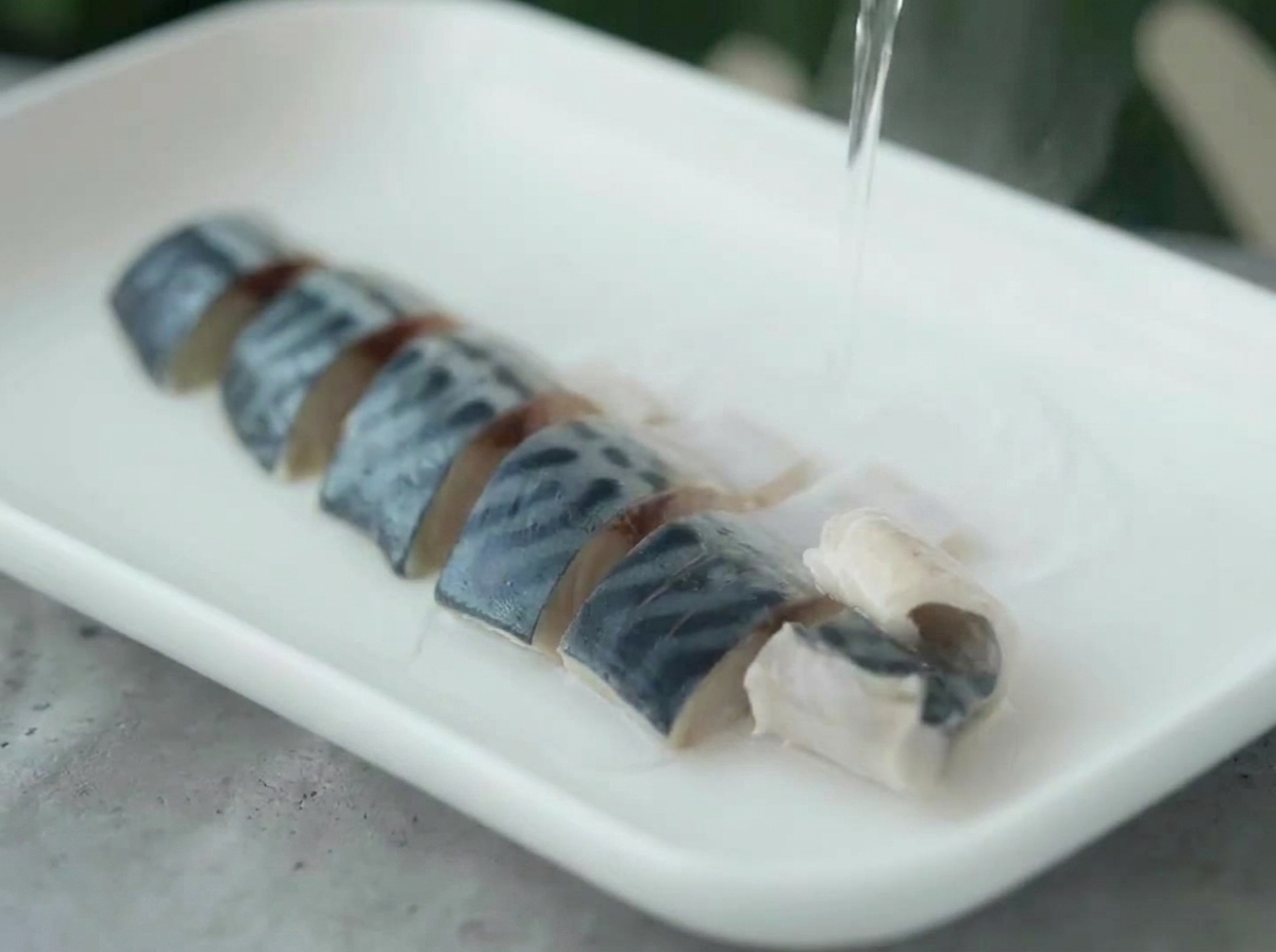 Acqua bollente versata da un bollitore su pezzi di sgombro disposti su un piatto bianco.
