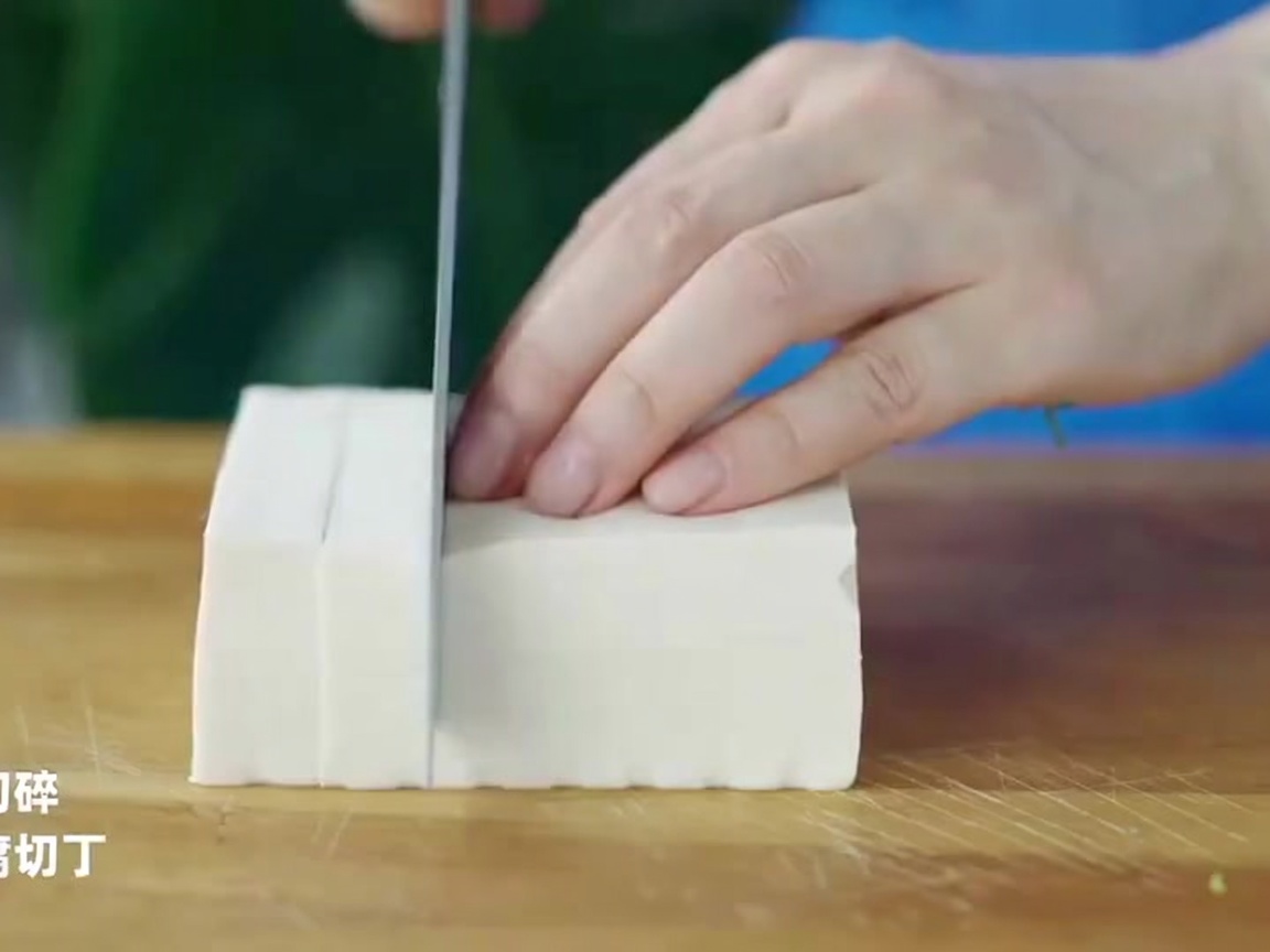 Un coltello da chef che taglia un panetto di tofu bianco a cubetti uniformi su una superficie di legno.