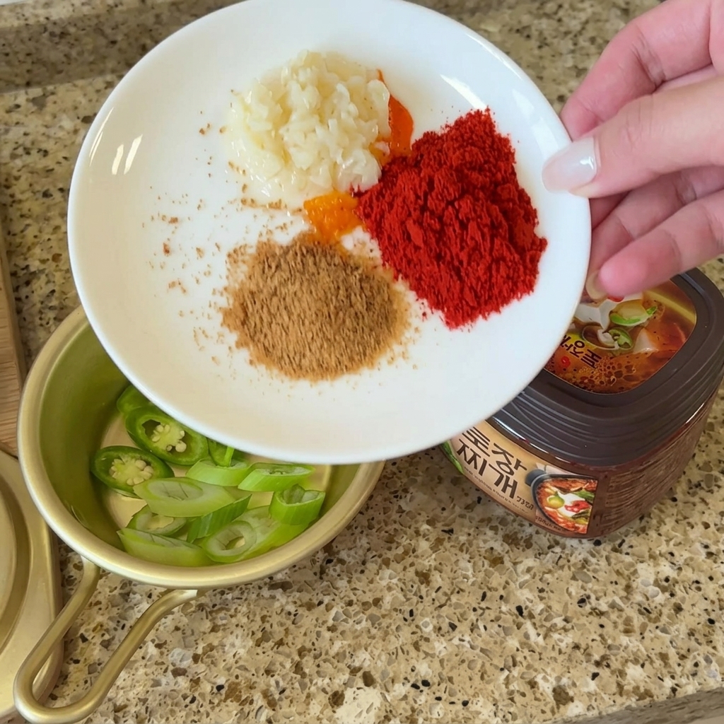 Un piatto bianco con porzioni di aglio tritato, peperoncino in polvere, brodo di manzo e polvere di perilla accanto a un contenitore di doenjang.