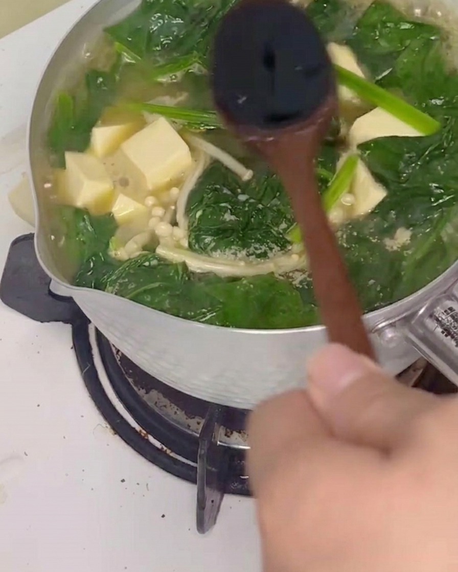 Salsa di soia aggiunta alla zuppa di spinaci e tofu che sobbolle, mescolata con un cucchiaio di legno.