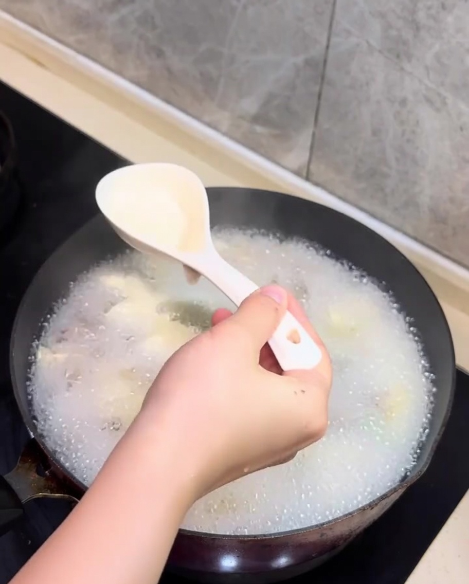Un cucchiaio che rimuove la schiuma bianca dalla superficie della zuppa di vongole e tofu in cottura.
