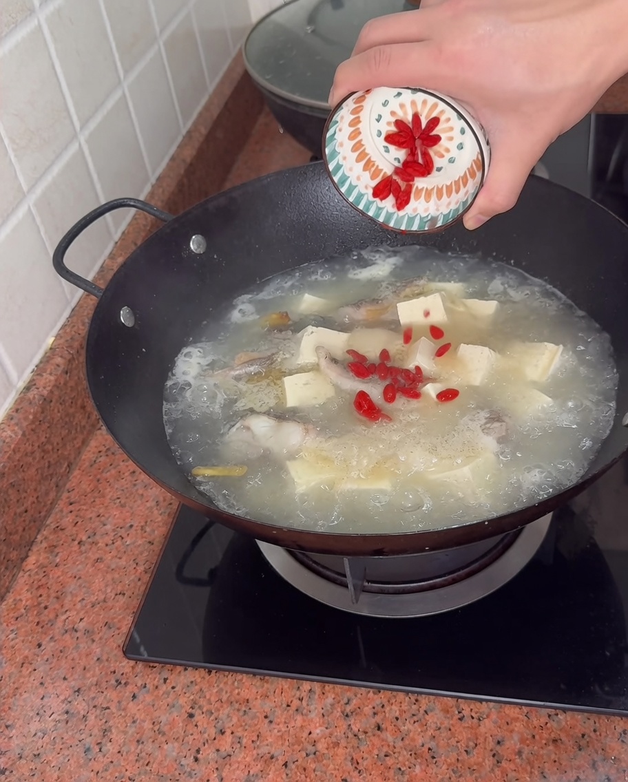 Versare le bacche di goji rosse da una piccola ciotola decorata in un wok ribollente di zuppa di pesce e tofu.