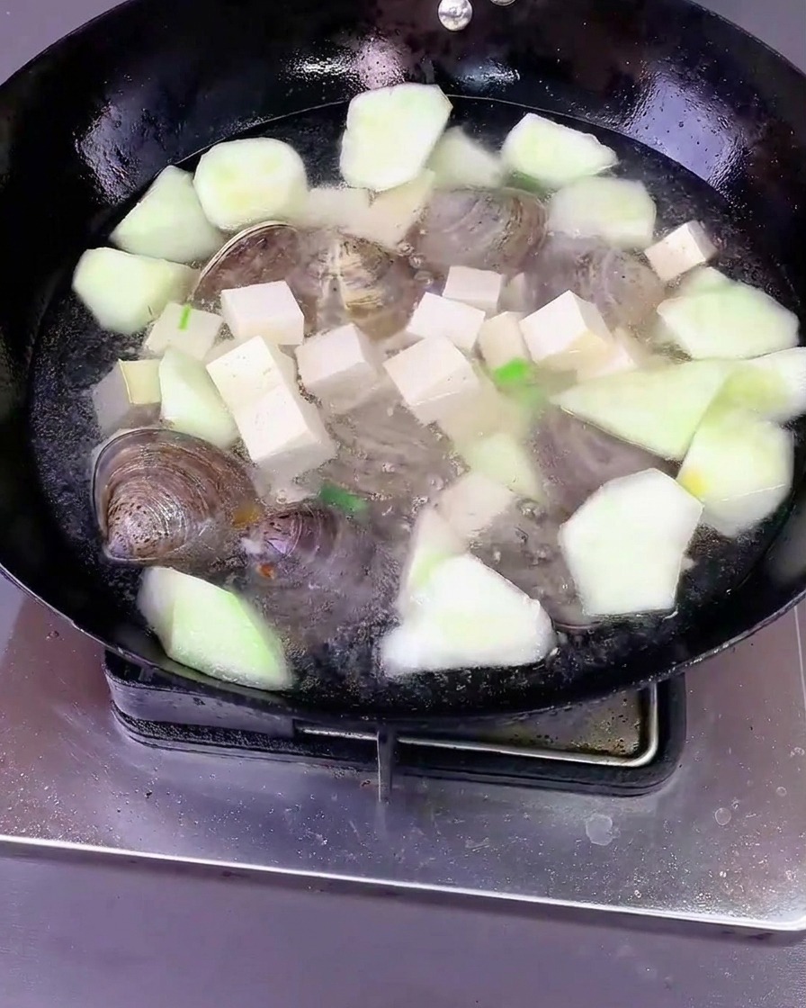 Acqua versata su luffa, tofu e vongole in un wok con aromatici e condimenti, coperto da un coperchio.