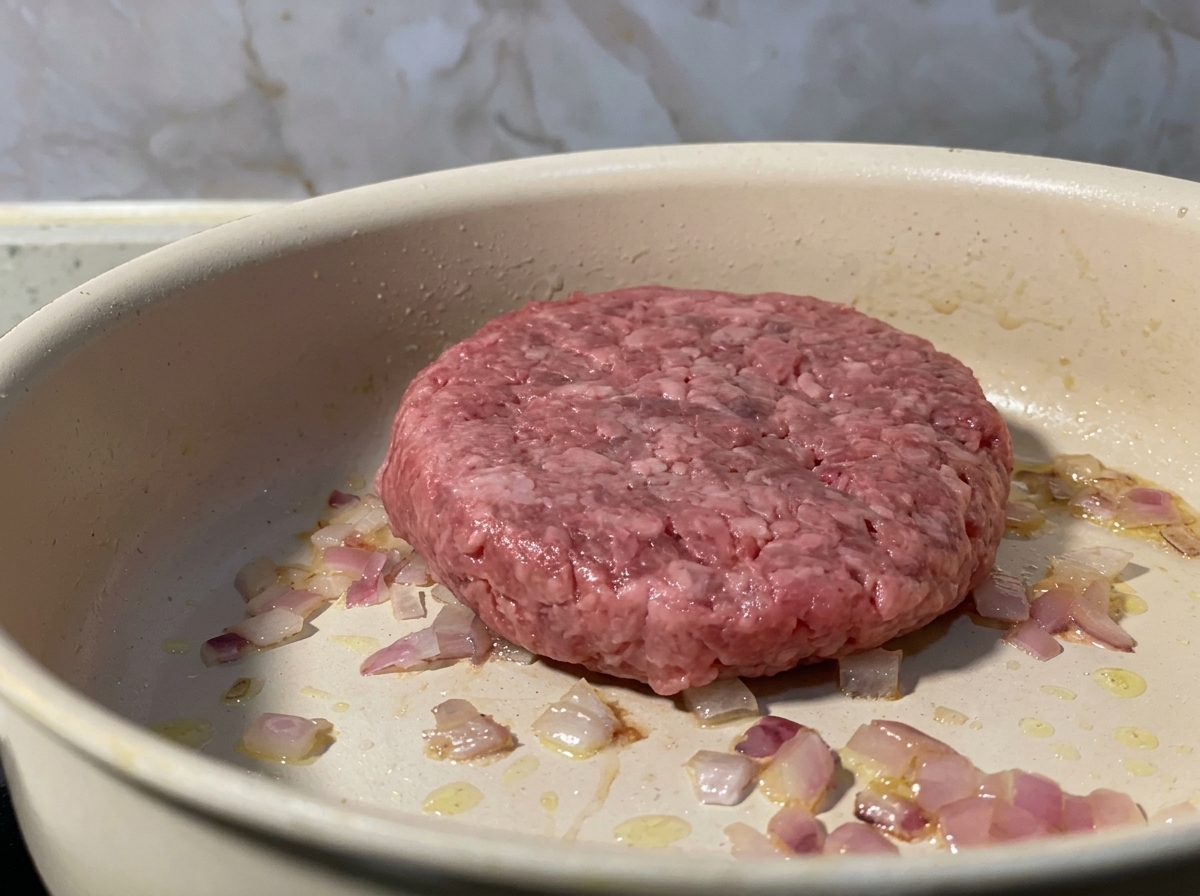 Un hamburger di manzo crudo in padella insieme alla cipolla rossa soffritta.