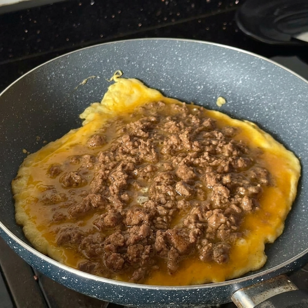 Una frittata rotonda che cuoce in una padella, ricoperta di manzo macinato cotto e speziato.