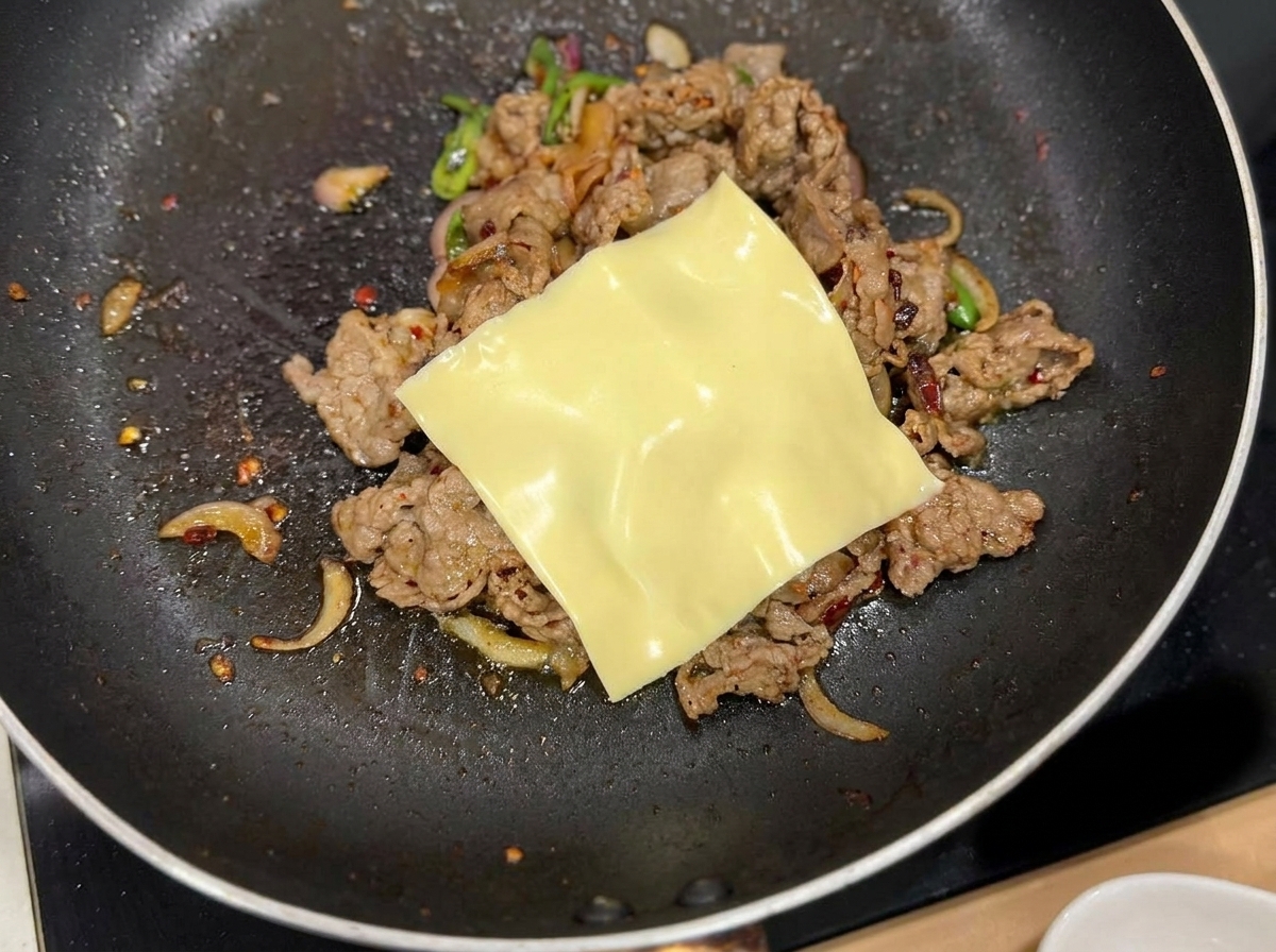 Una fetta di formaggio a pasta gialla che si scioglie sul composto cotto di manzo e cipolle in padella.