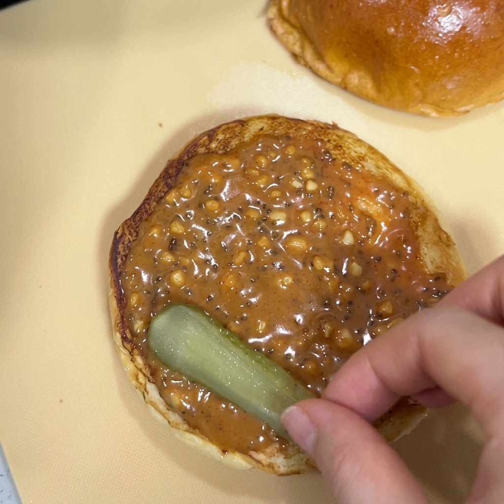 Una mano che posiziona una fetta di cetriolino su un panino coperto di densa salsa di arachidi.