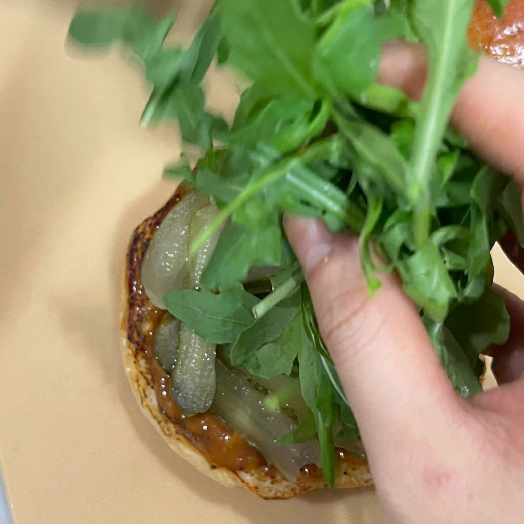 Una mano che accumula foglie di rucola fresca sui cetriolini alla base del panino.