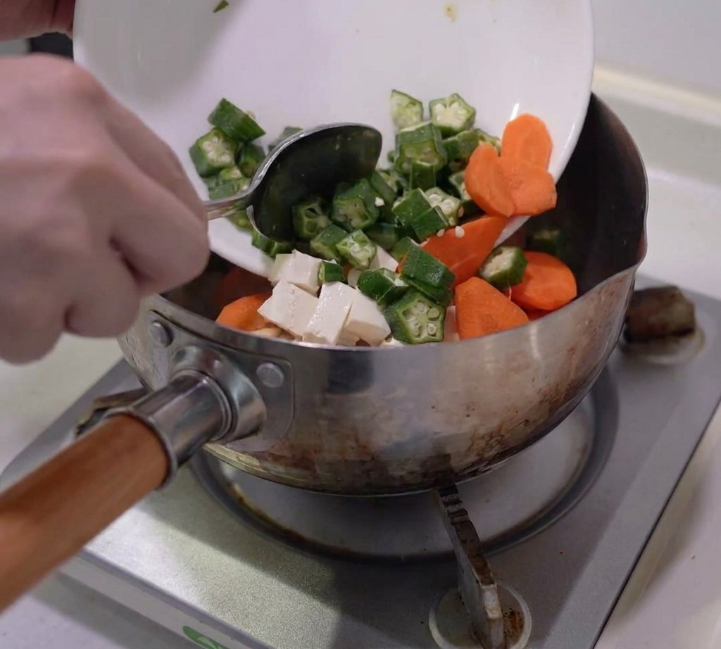Okra, carote, funghi e tofu preparati vengono aggiunti in una pentola dacciaio.