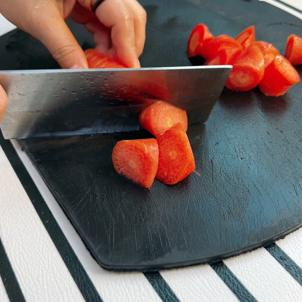 Una mano usa una mannaia per tagliare una carota in pezzi irregolari su un tagliere nero.