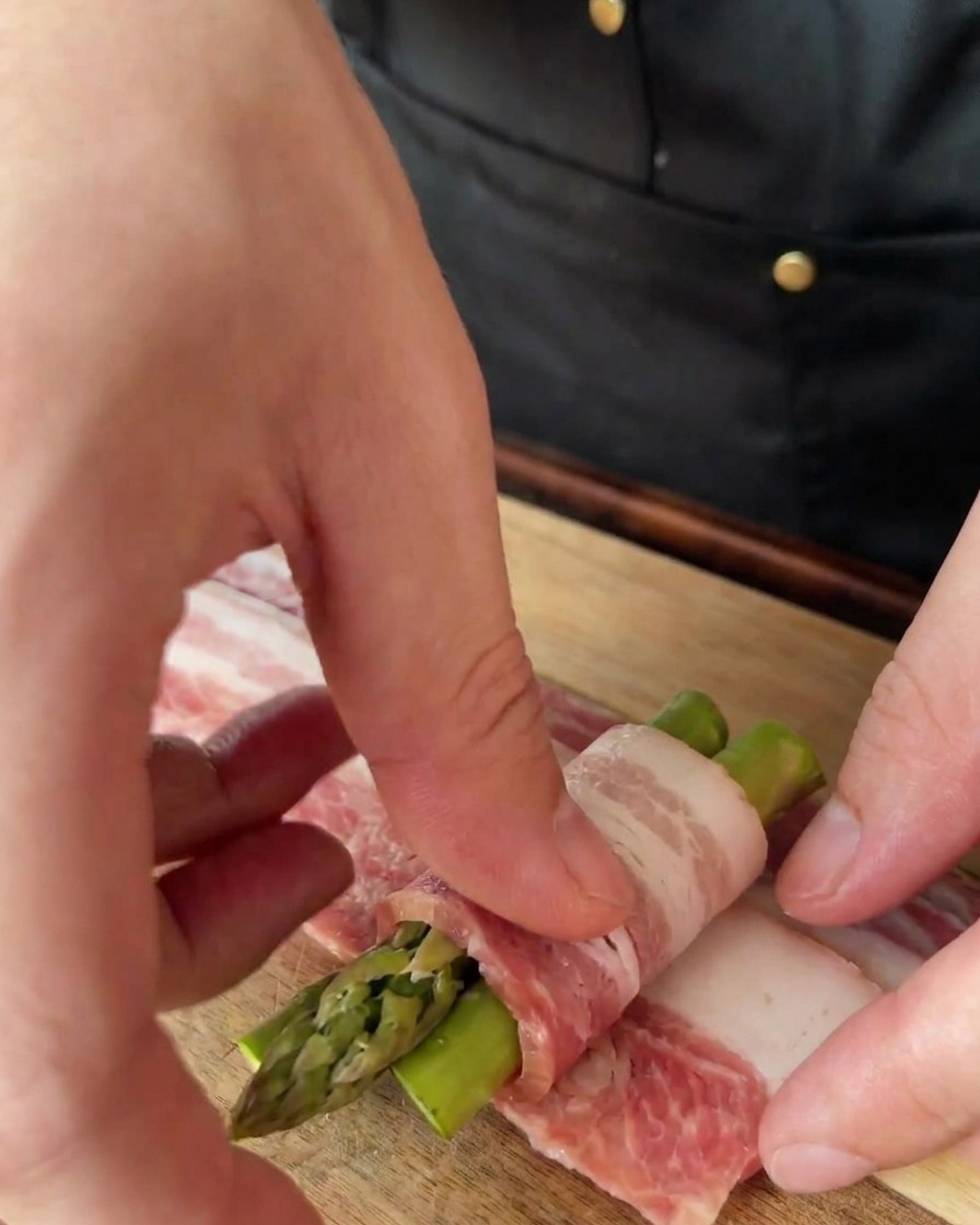 Mani che avvolgono una fetta sottile di pancetta attorno a un mazzetto di tre asparagi.