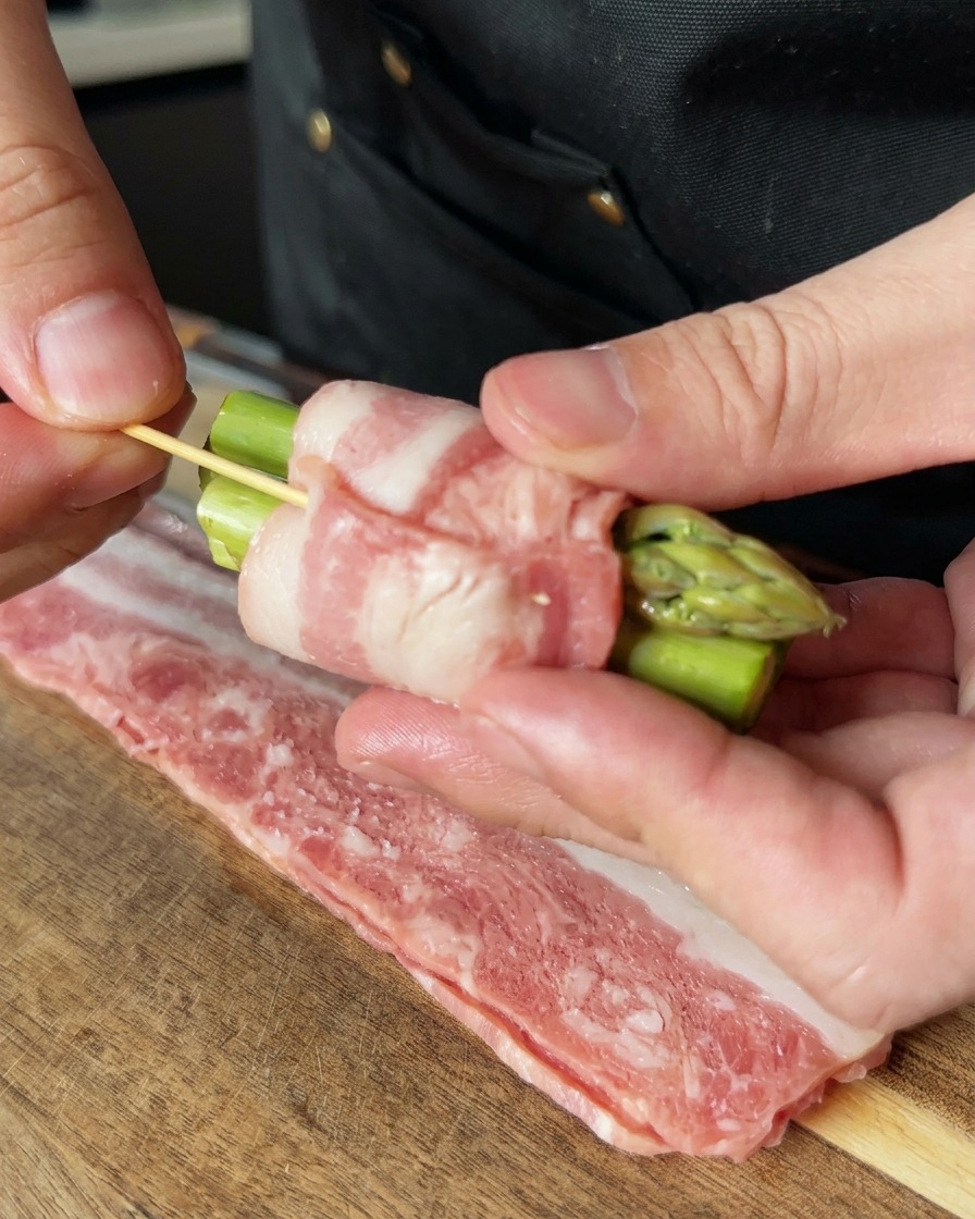 Una mano usa uno stuzzicadenti di legno per fissare un involtino di asparagi e pancetta.